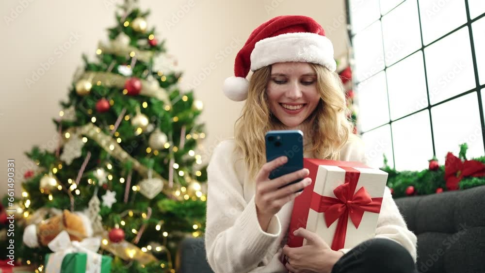 Young blonde woman using smartphone holding christmas gift at home Young blonde woman using smartphone holding christmas gift at home