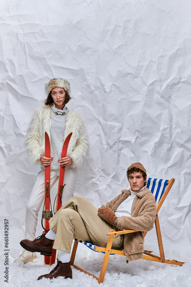 winter fashion and leisure, asian woman with skis near trendy man in deck chair in snowy studio winter fashion and leisure, asian woman with skis near trendy man in deck chair in snowy studio