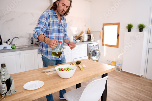 Handsome Caucasian bearded man pouring some olive oil into a white bowl with fresh healthy salad.