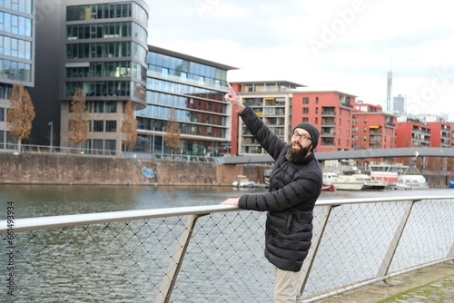 young bearded man in coat and hat stands on shore river Main, points to View of modern residential buildings, winter walks in Frankfurt, Gutleitviertel district, boats and yachts moored on banks photo