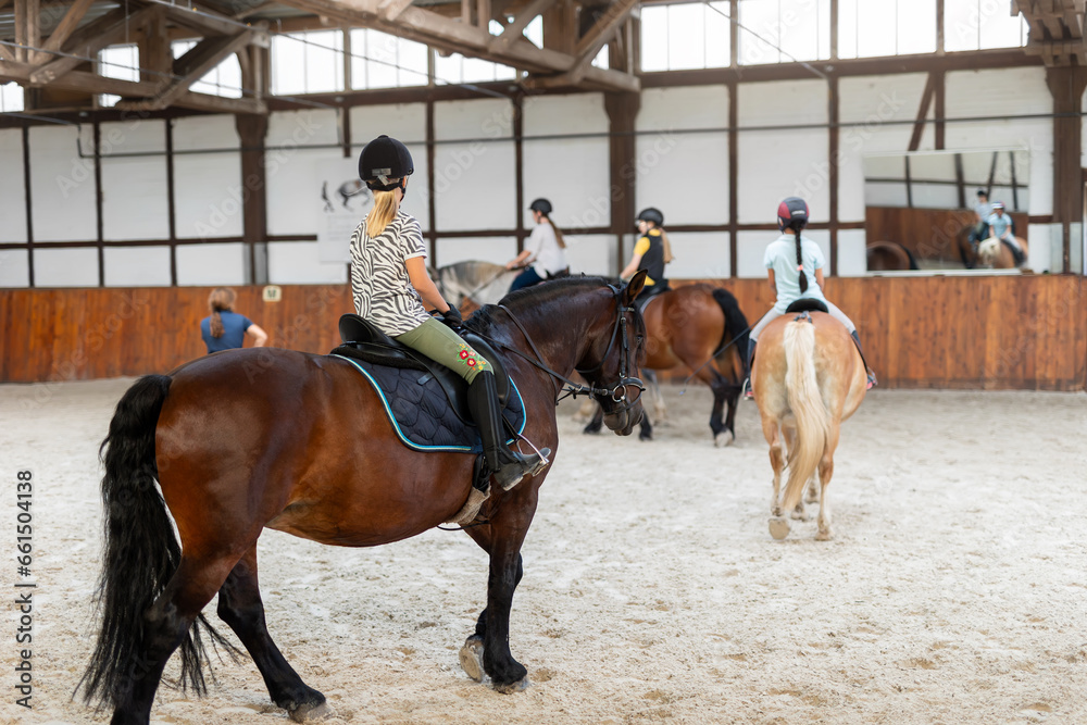Horse riding school. Little children girls at group training equestrian lessons in indoor ranch horse riding hall. Cute little beginner blond girl kid in helmet sitting on brown horse horseback Horse riding school. Little children girls at group training equestrian lessons in indoor ranch horse riding hall. Cute little beginner blond girl kid in helmet sitting on brown horse horseback