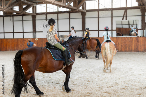 Horse riding school. Little children girls at group training equestrian lessons in indoor ranch horse riding hall. Cute little beginner blond girl kid in helmet sitting on brown horse horseback