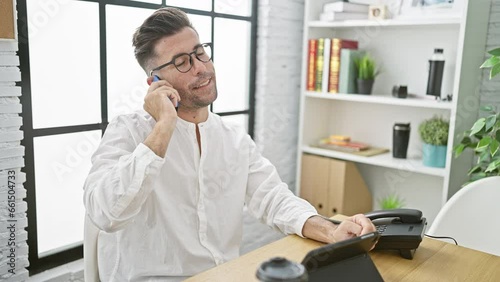 Wallpaper Mural Confident young hispanic businessman joyfully engaged in a conversation on his smartphone while working at the office desk, amidst a professional indoor setting Torontodigital.ca