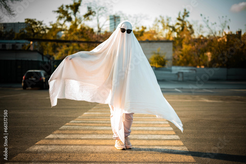 Fototapeta Naklejka Na Ścianę i Meble -  Celebrating halloween. Ghost Challenge 2021. A man dressed as a ghost from a sheet and sunglasses crosses the road along the crosswalk. Spooky season.