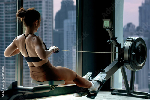 Fitness girl workout on indoor rower at the gym, doing exercises on rowing machine near the window,  view of skyscrapers in the background, woman exercising in fitness club