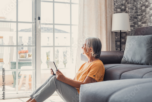 Smiling middle-aged Caucasian woman sit on couch in living room browsing wireless Internet on tablet, happy modern senior female relax on ground at home using pad device, elderly technology concept.