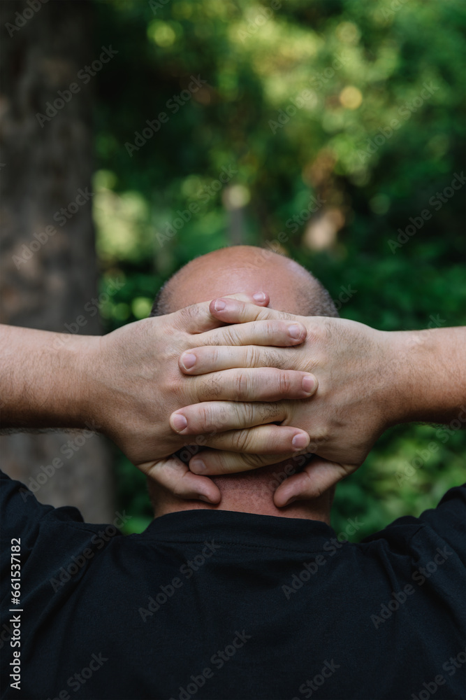 Fototapeta premium View from behind of mature man with little hair or alopecia with his hands on the nape of his neck. Outdoor vertical photography outdoors with natural light. Concept of reflection and observation 