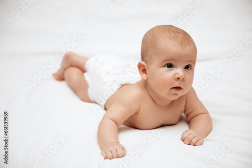 A newborn baby in a diaper is lying on stomach on a white background and looking to the side.