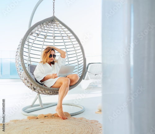 Beautiful middle-aged woman sitting in a comfortable hanging chair on the open house terrace with a modern laptop and browsing in internet doing shopping online. Modern technology and home lifestyle.