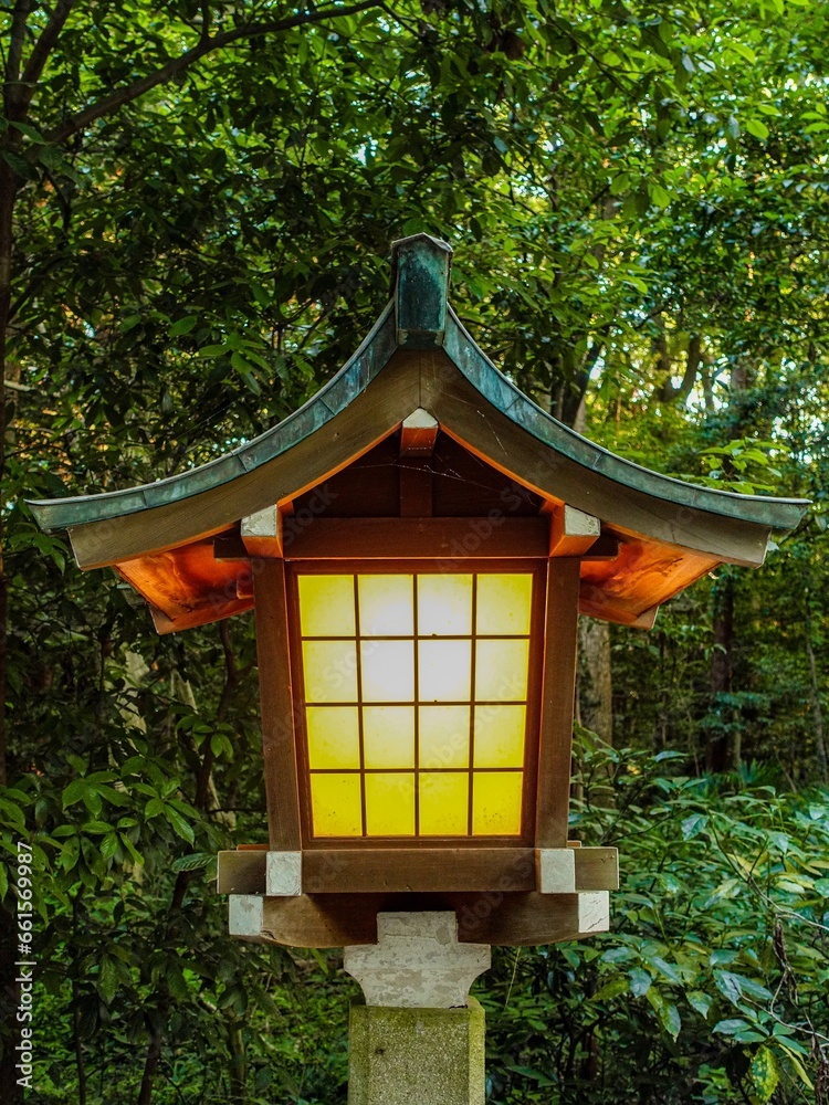 Closeup of old, traditional Japanese lantern with curved roof, glowing orange light during the evening, with green forest in the background. Closeup of old, traditional Japanese lantern with curved roof, glowing orange light during the evening, with green forest in the background.