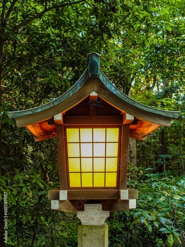 Closeup of old, traditional Japanese lantern with curved roof, glowing orange light during the evening, with green forest in the background. © Kosma Sobol