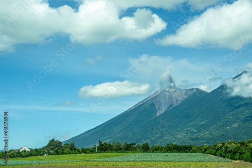 Guatemala, La Antigua - July 20, 2023: Agriculture at base of erupting De fuego volcano above and SW of the town under blue cloudscape. Green jungle in front photo