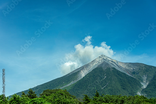 Guatemala, La Antigua - July 20, 2023: Smoke plume erupts from De fuego volcano above and SW of the town under blue cloudscape. Green jungle in front