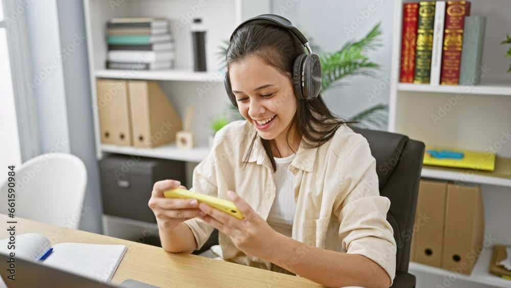 Smiling young hispanic woman worker enjoying video on her smartphone, using headphones in a bustling office, exhibiting a beautiful mix of elegance and professional success. Smiling young hispanic woman worker enjoying video on her smartphone, using headphones in a bustling office, exhibiting a beautiful mix of elegance and professional success.