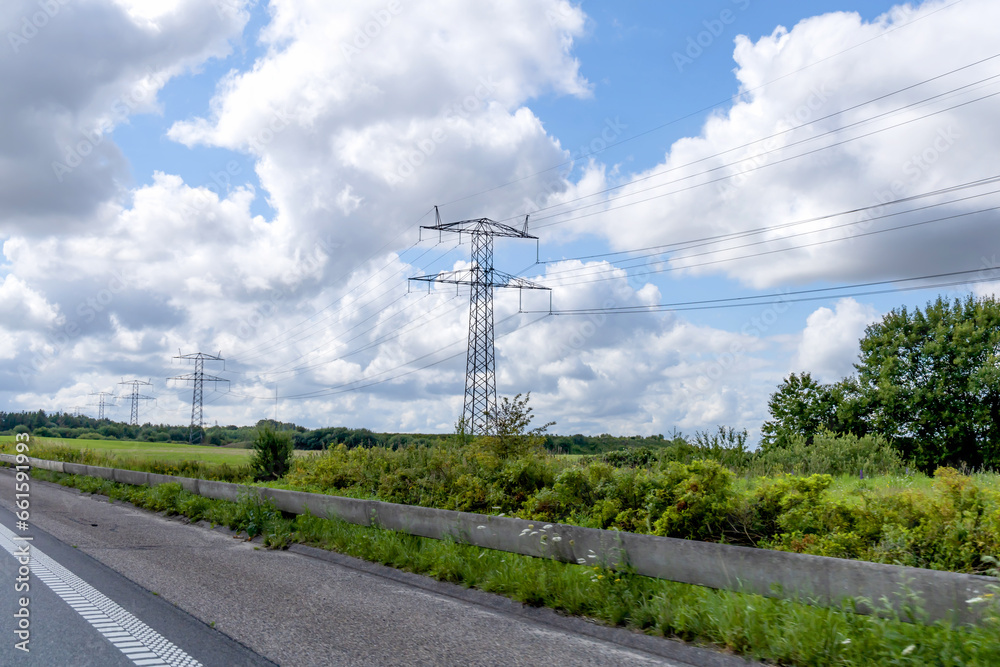  High voltage power line pylons, electrical tower on a green field with blue sky. Highway with cars on a cloudy summer day.