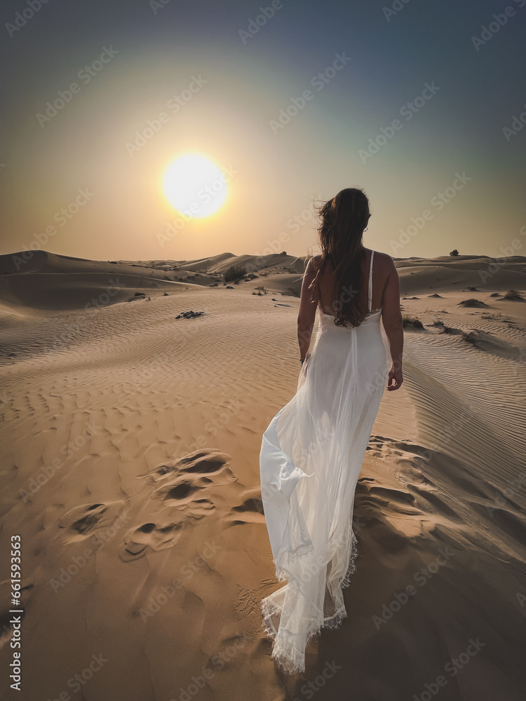 view from the back of a slender tanned girl standing in the middle of the desert under the scorching sun in a white sexy dress blowing in the wind