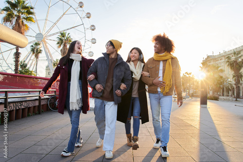 Group of cheerful multi-ethnic friends enjoying winter vacations outdoor. Happy laughing young people together holding arm at amusement park. Multicultural Generation z walking having fun at sunset.