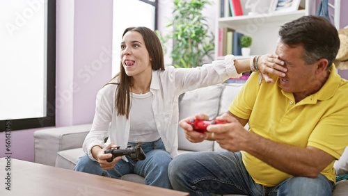 Hispanic father and daughter enjoy playing video game on the cozy home sofa, covering eyes and hands in laugh-filled skulduggery photo