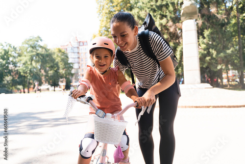 Mother learning child girl to ride a bike in the park in the summer. Happy quality family time together. Empowering, essential physical skills for kids.