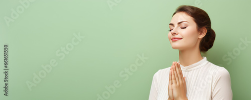Young peaceful woman with closed eyes praying with folded hands in prayer gesture, blessing. Isolated on flat pastel green background with copy space, banner template. photo