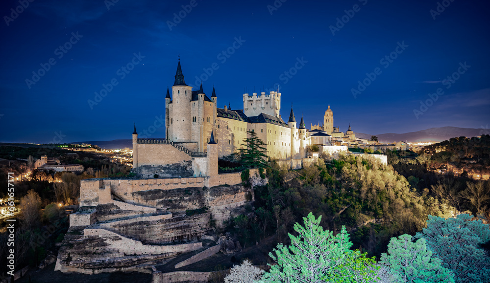 Castillo medieval europeo sobre una colina por la noche, con algunas estrellas, vegetación en el primer plano y una ciudad también medieval al fondo, desde la ciudad de Segovia, España.
