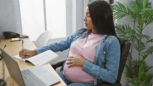 Expecting young hispanic businesswoman, task-juggling in her profession, sensitively balancing pregnancy and work - note-taking on her laptop in a relaxed office photo