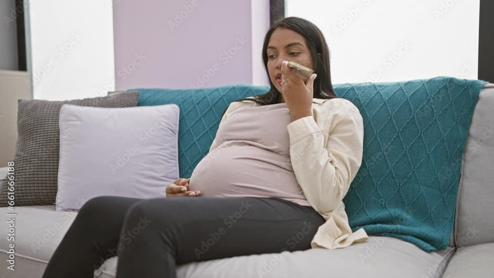 Expecting young woman, comfortably relaxing on her living room sofa, tenderly touching her pregnant belly while send, speaks a heartfelt voice message via her smartphone at home Expecting young woman, comfortably relaxing on her living room sofa, tenderly touching her pregnant belly while send, speaks a heartfelt voice message via her smartphone at home