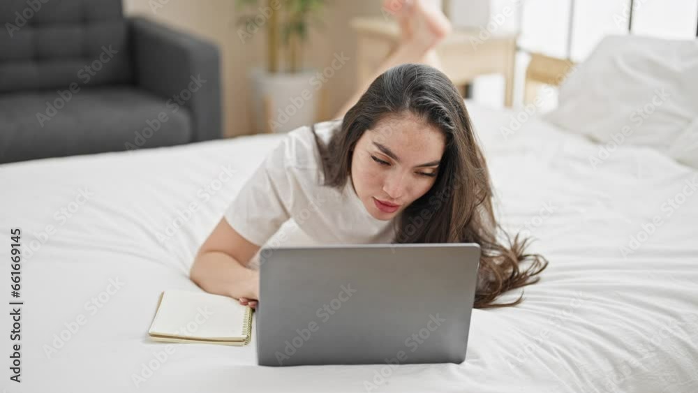 Young beautiful hispanic woman using laptop taking notes at bedroom