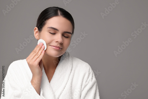 Young woman cleaning her face with cotton pad on grey background. Space for text