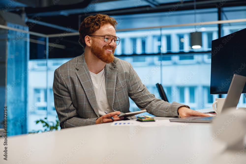 Fototapeta premium Handsome businessman working on laptop and making notes while sitting desk on office background