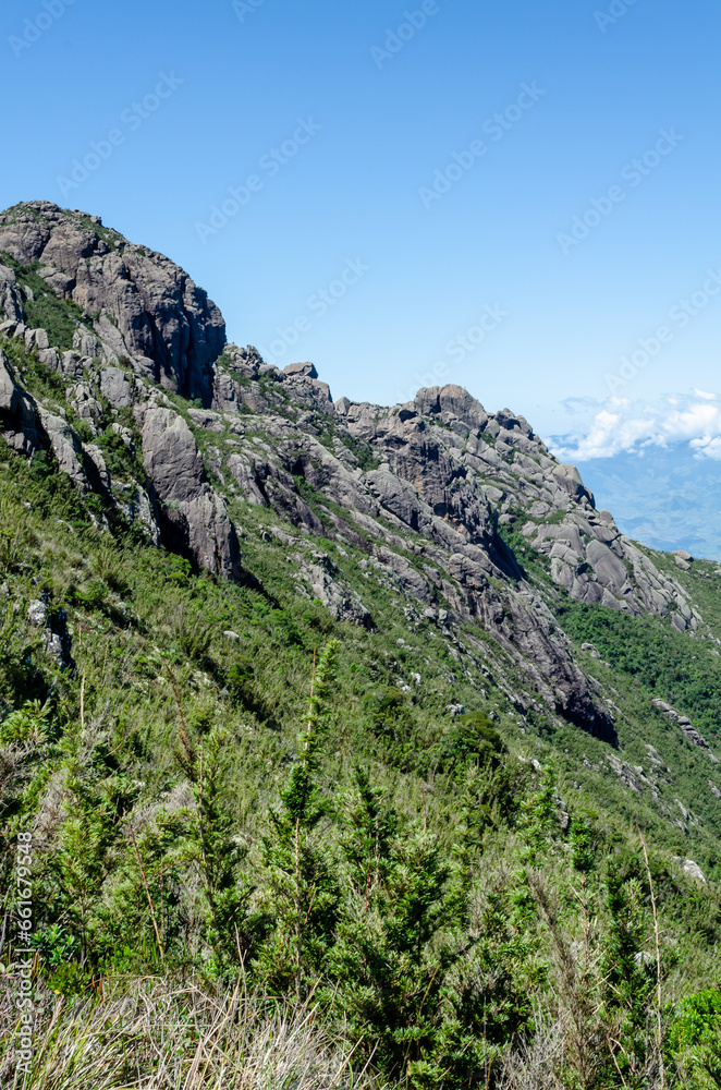 mountainous landscape with a clear blue sky. The mountain is covered in green vegetation and has a rocky cliff face.