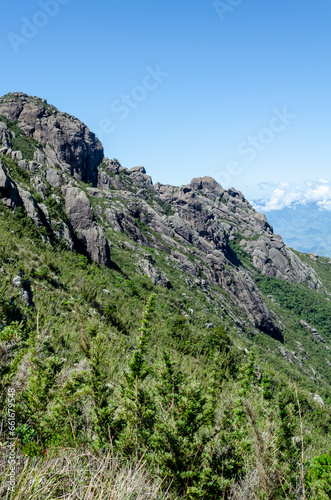 mountainous landscape with a clear blue sky. The mountain is covered in green vegetation and has a rocky cliff face.