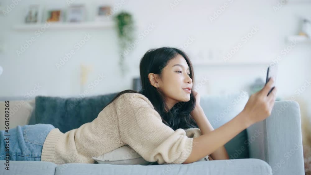 Happy young asian woman relaxing at home. Female smile lying on sofa and holding mobile smartphone. Girl using video call to friend Happy young asian woman relaxing at home. Female smile lying on sofa and holding mobile smartphone. Girl using video call to friend