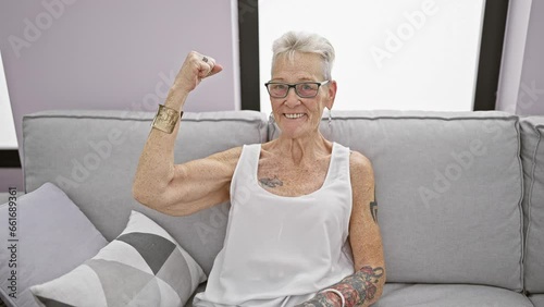 Confident grey-haired senior woman joyfully flexing her biceps on the living room sofa at home, sporting a big cheerful smile and a cool, positive attitude
