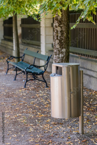 Metal trash can beside walkway in public park with blurred background.