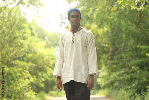 An Indian Boy wearing Khadi Kurta and a Bandana or neck gaiter standing on the road with green trees and vegetation in the background, smiling while wearing specs