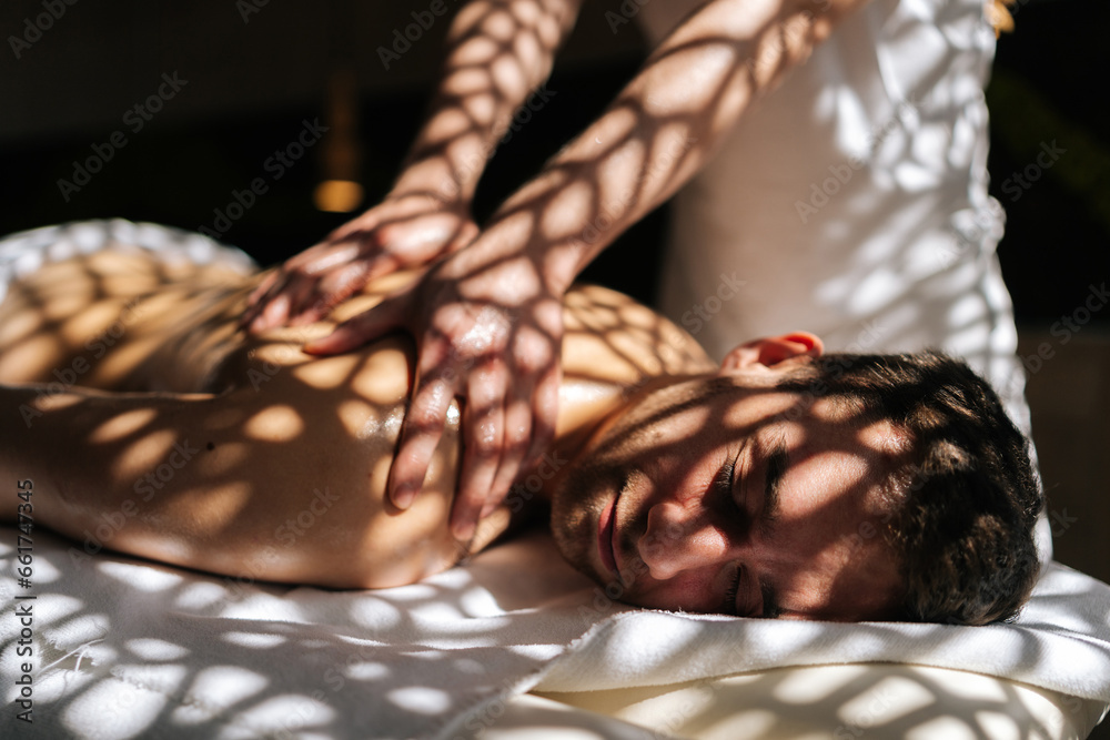 Closeup face of happy man getting professional back massage from masseur male in spa salon lying on massage table with closed eyes in sunlight and beautiful shadows. Concept of healthy lifestyle. Closeup face of happy man getting professional back massage from masseur male in spa salon lying on massage table with closed eyes in sunlight and beautiful shadows. Concept of healthy lifestyle.