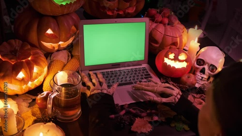 Top view on laptop with chromakey screen and bony hands using keyboard near many Halloween flashing orange pumpkins, candles, glass of beer, skull. Advertising platform. Happy Halloween background. photo