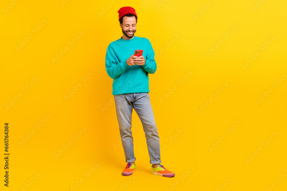 Full length photo of satisfied man dressed turquoise pullover look at smartphone read notification isolated on yellow color background Full length photo of satisfied man dressed turquoise pullover look at smartphone read notification isolated on yellow color background