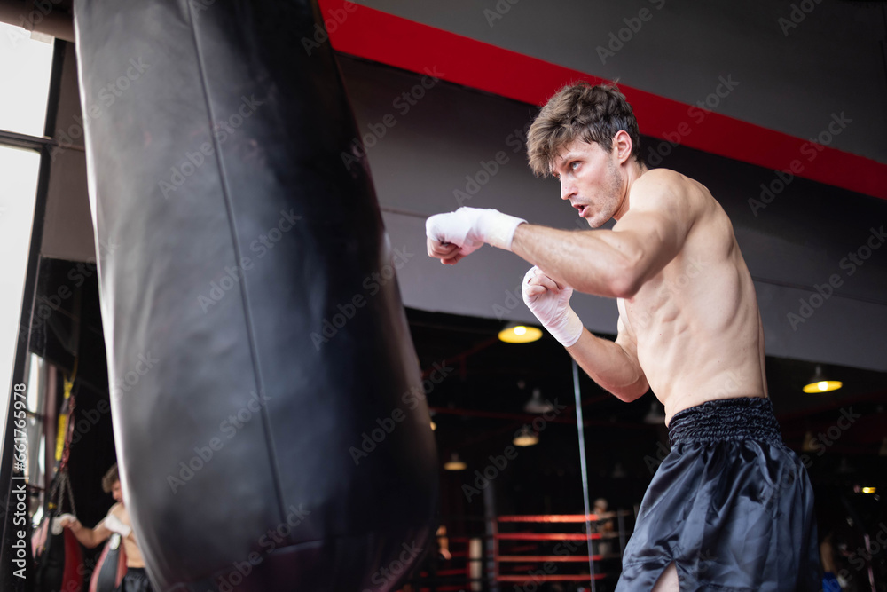 active Caucasian professional athlete, dressed in sportswear and boxing gloves, is engaging in a workout session by striking a punching bag or boxing sandbag at a gym or fitness club.
