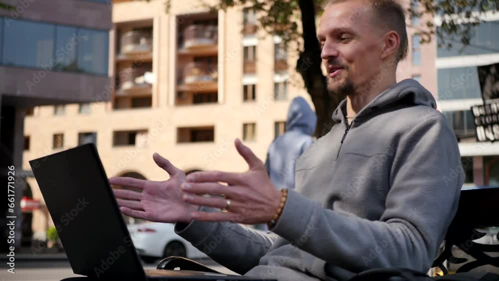 Caucasian young stylish handsome man sitting on a bench and talking on a video call using a laptop. Adult handsome blond man using laptop computer outdoors in the city and gesturing with his hands Caucasian young stylish handsome man sitting on a bench and talking on a video call using a laptop. Adult handsome blond man using laptop computer outdoors in the city and gesturing with his hands