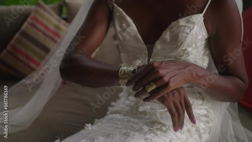 Non-binary black bride in fashionable white wedding dress, gold jewelry rings puts on brass vintage bracelet with dark gem on hand. Androgynous bipoc person of colour prepares for wedding. photo