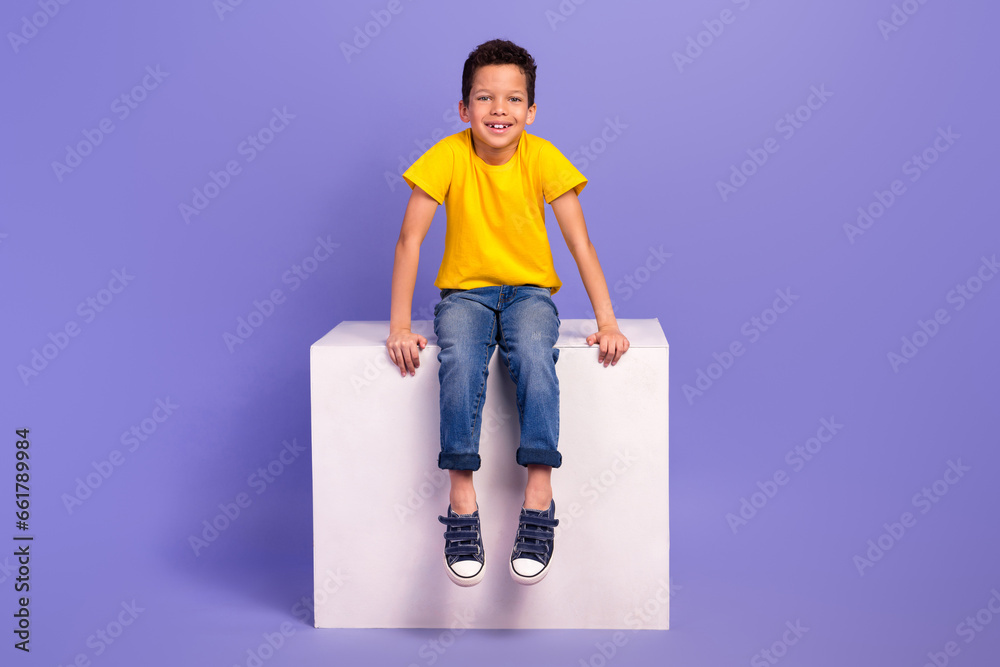Full body photo of charming young boy sit white cube shopping promo dressed yellow outfit isolated on violet color background - obrazy, fototapety, plakaty 