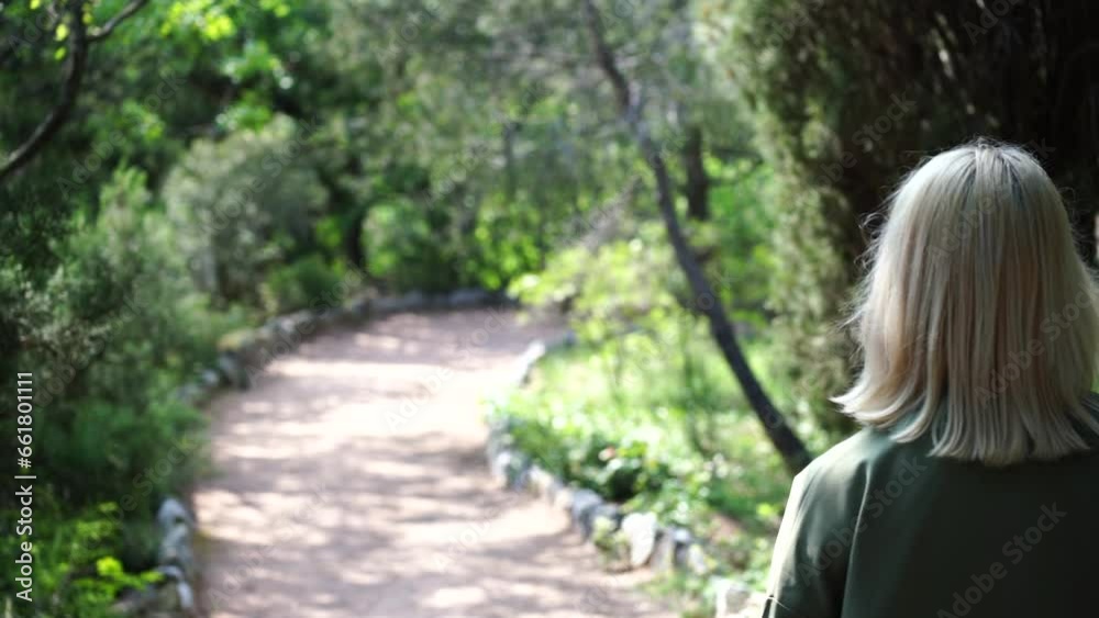 Blonde walking in the park. Happy young woman wearing. Happy young woman in a green jacket walks forward in the park, rear view, slow motion Blonde walking in the park. Happy young woman wearing. Happy young woman in a green jacket walks forward in the park, rear view, slow motion