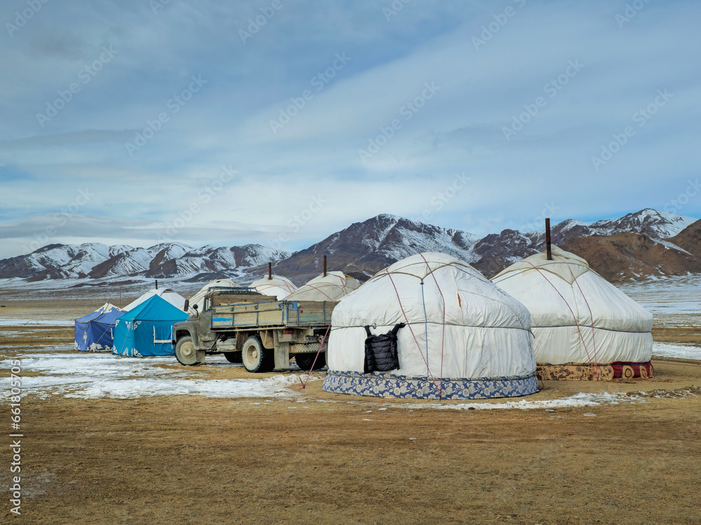 Mongolian yurt camp in the winter steppe. An old truck stands next to the yurts against the backdrop of snow-capped mountains.