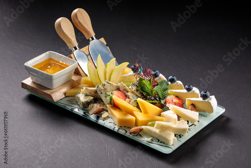 
Snack board with different types of cheese, fruits, berries and knives on a black background