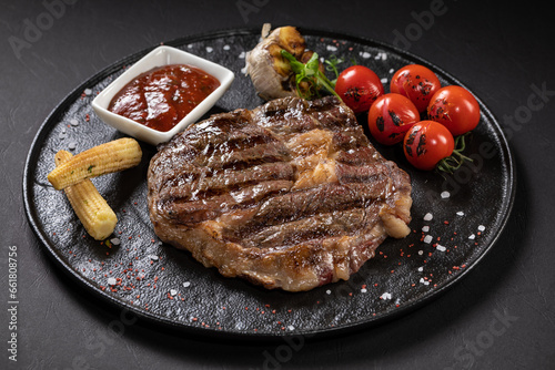 BBQ beef steak with sauce on a black plate with vegetables, tomatoes and corn. Black background. Restaurant supply.