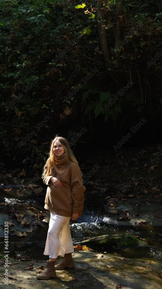 a beautiful pregnant woman in a sweater stands on a stone on the background of a mountain river on a sunny day and strokes her belly and looks at the camera. vertical video a beautiful pregnant woman in a sweater stands on a stone on the background of a mountain river on a sunny day and strokes her belly and looks at the camera. vertical video
