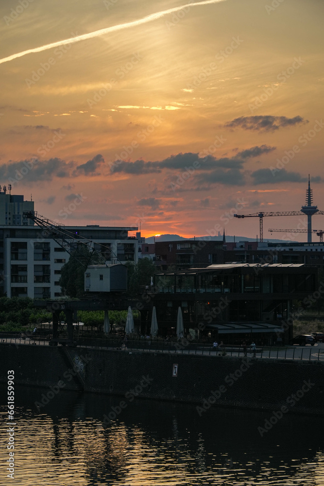 buildings and a crane next to a river. city buildings, the view tower Europaturm and the taunus mountains in the backround. On top the sun sets behind the mountains