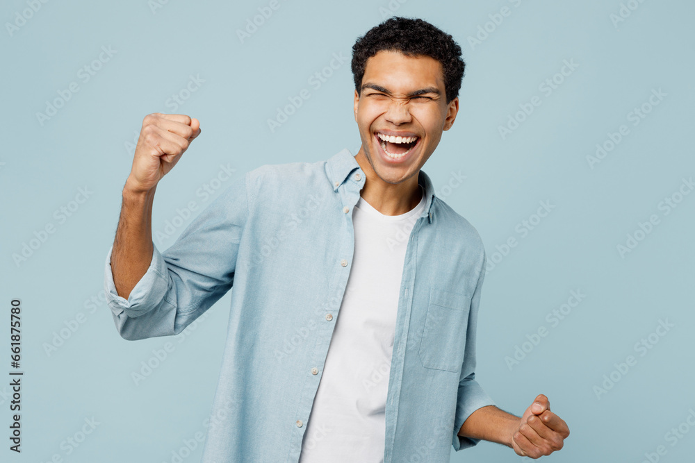Young man of African American ethnicity wear shirt white t-shirt casual clothes doing winner gesture celebrate clench fists say yes isolated on plain pastel light blue cyan background studio portrait. Young man of African American ethnicity wear shirt white t-shirt casual clothes doing winner gesture celebrate clench fists say yes isolated on plain pastel light blue cyan background studio portrait.